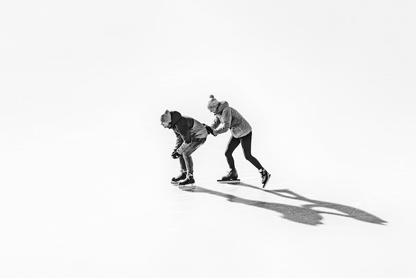 Two people in snowclothes and ice skates; one is pushing the other forward